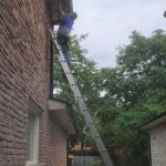 A technicians standing on a tall ladder while repairing eavestrough on a roof.